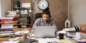 Student writing thesis at a busy desk with books.